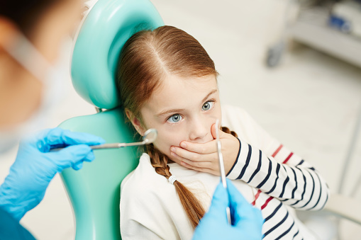 Scared little girl covering her mouth by hand while looking at dental tools for oral check-up held by dentist